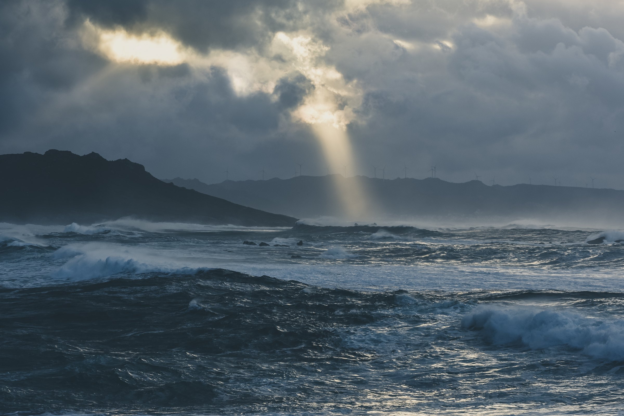 Rough grey sea under dark clouds with a shaft of light breaking through over a distant headland
