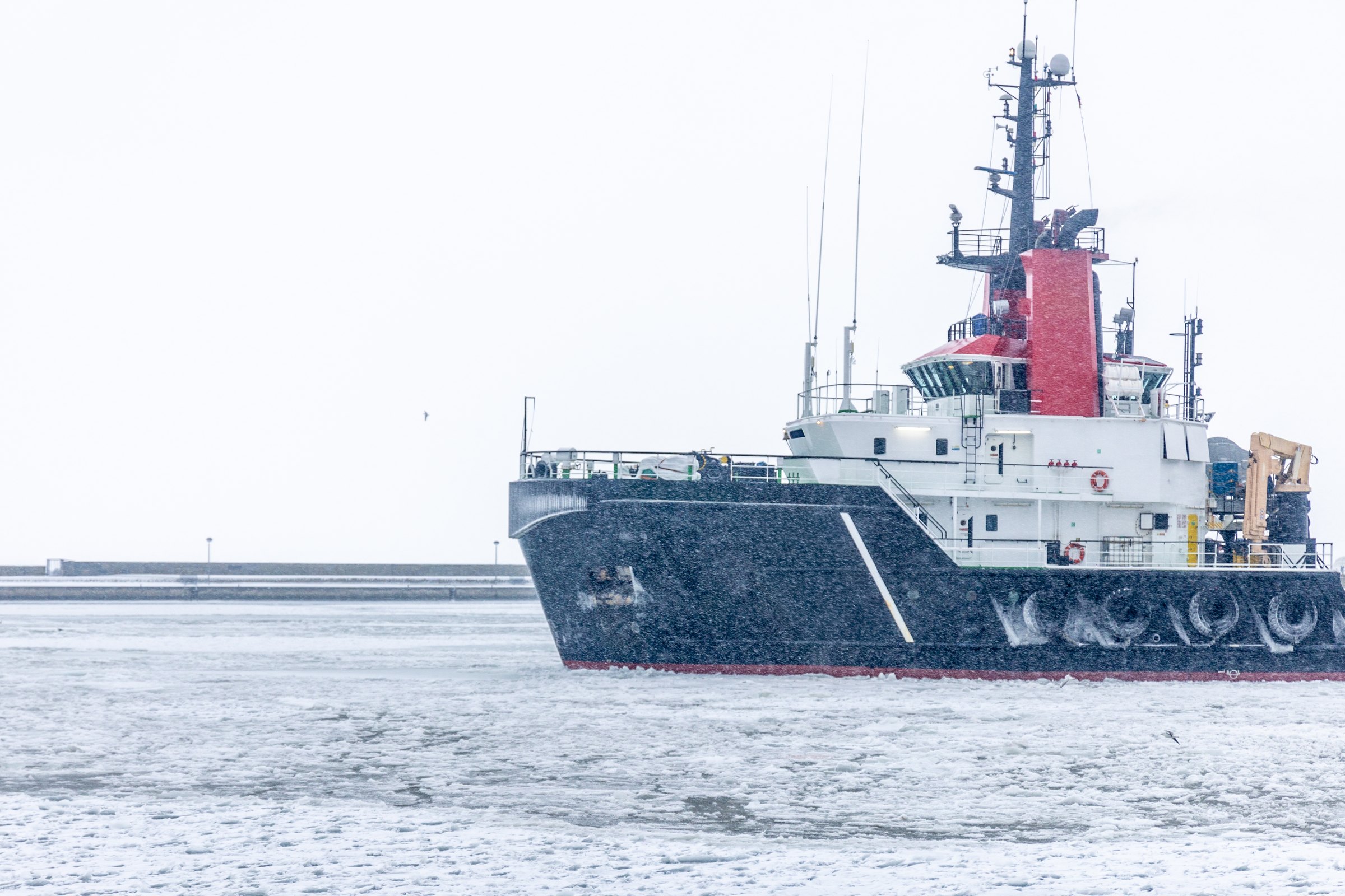 A tug-type work vessel moving through ice-covered harbour water in falling snow