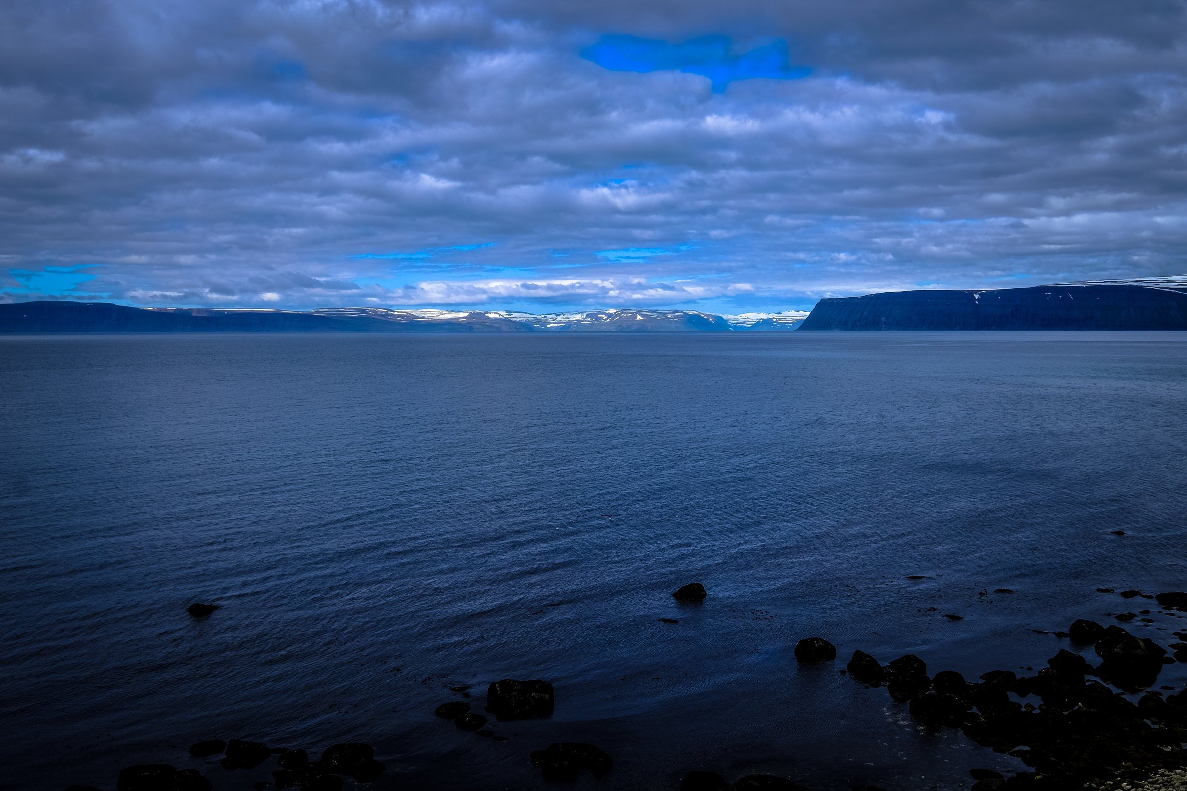 Dark blue open water at dusk with a distant line of snow-capped coast under heavy cloud