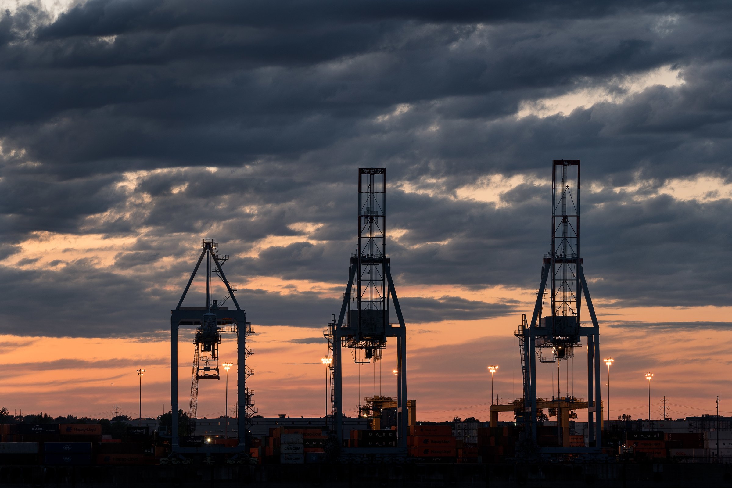 Silhouettes of three container gantry cranes against a cloudy evening sky