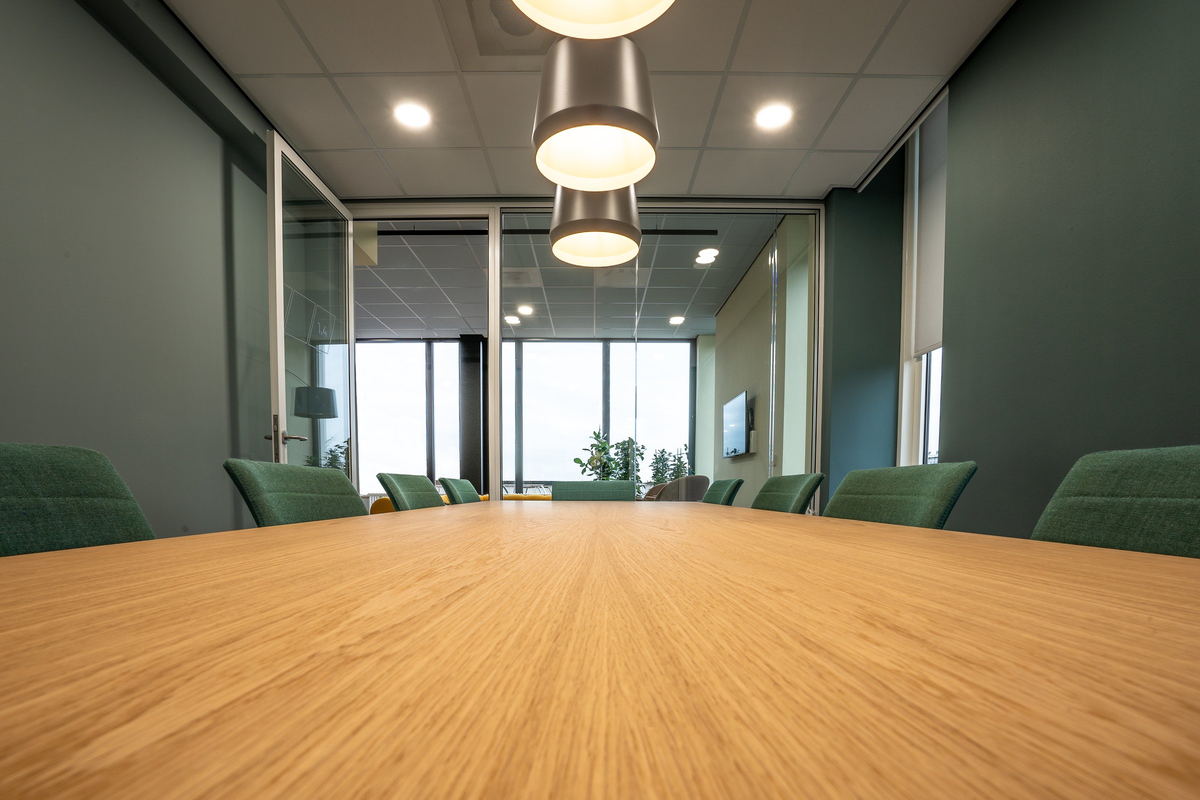 Low view across an empty wooden boardroom table with green upholstered chairs and pendant lamps