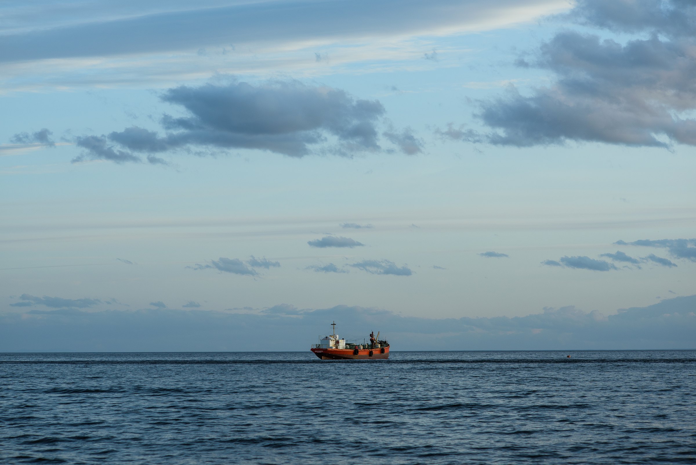 Small orange work vessel on the horizon of a calm, open sea under a pale blue sky