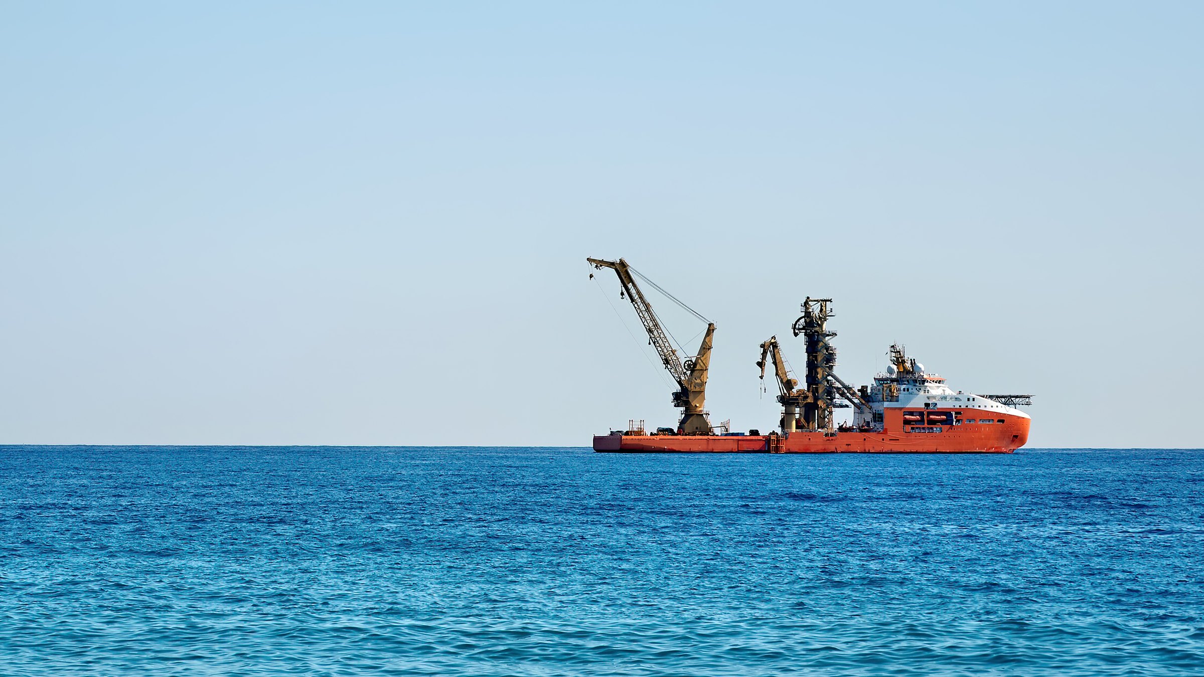 Red offshore construction vessel with two large cranes and a tower working far out on a bright blue sea