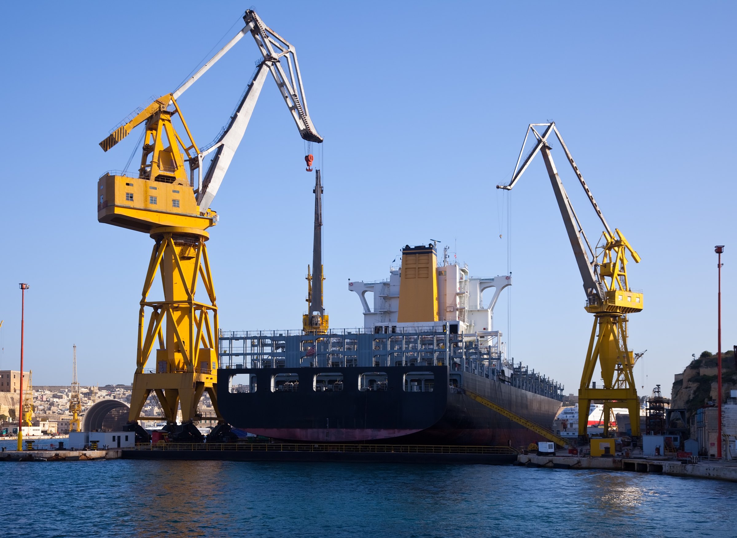 A large ship in a dry dock between two yellow dockside cranes on a clear day